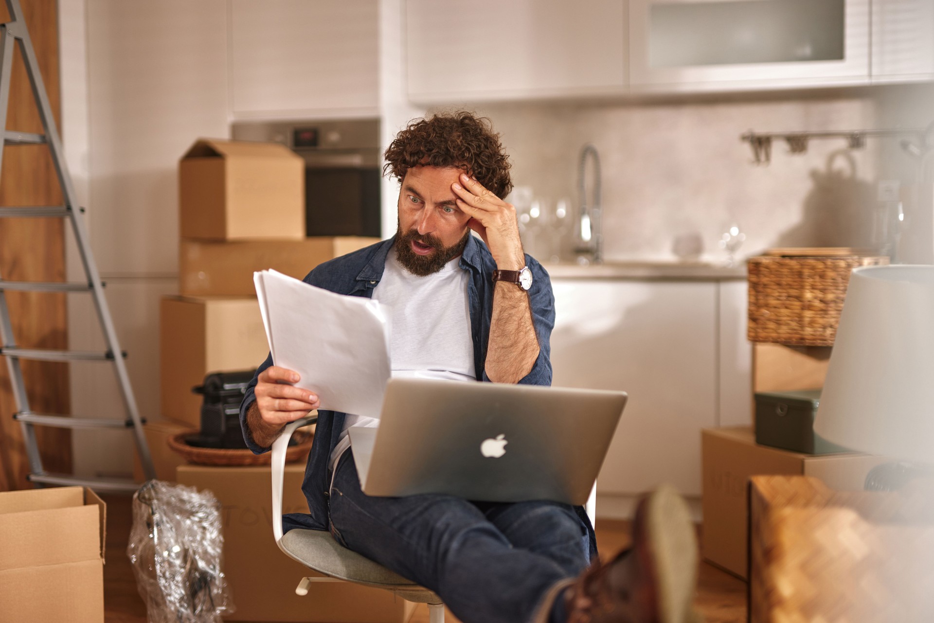 Stressed man reading documents and using laptop while moving into new home